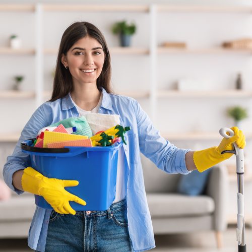 Portrait of smiling millennial lady holding bucket with cleaning supplies and mop, posing and looking at camera standing in living room. Professional cleaning service specialist wearing rubber gloves
