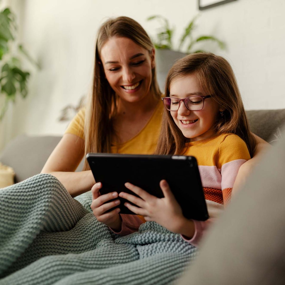Smiling young Caucasian mother and small preschooler daughter sit relax on couch in living room using tablet. Happy mom or nanny and little girl child rest on sofa watch video on tablet.
